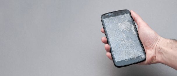 Hand holding mobile phone with broken screen on grey background. banner with copy space - Photo, image