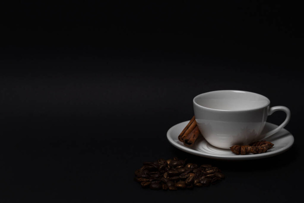 Empty white cup and saucer, coffee beans, cinnamon sticks, anise on black background - Photo, Image