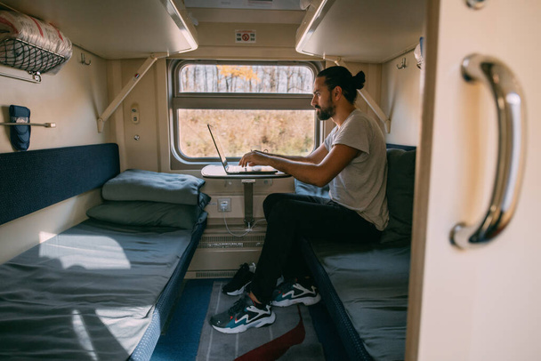 Hombre con portátil en el tren. Retrato. Joven chico guapo trabaja remotamente en un viaje en tren solo en un compartimiento de tren por la ventana - Foto, imagen