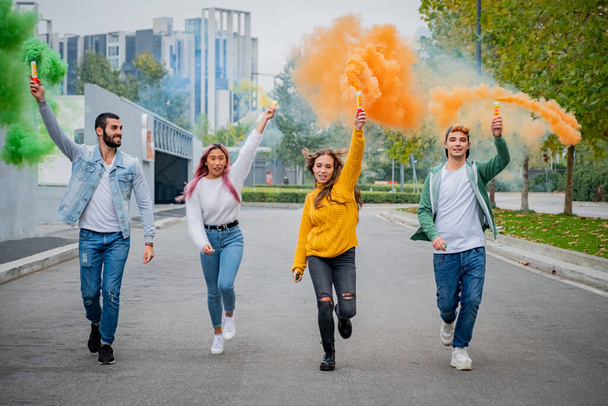 Красиві молоді хлопці стоять на вулиці і запалюють кольорові димові бомби - Щасливі друзі розважаються на вулиці з різнокольоровими димовими бомбами - Молоді студенти тисячоліття святкують весняні канікули разом
 - Фото, зображення
