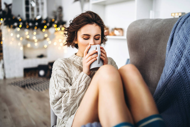 Pretty and happy brunette sitting in warm sweater on the chair, holding cup of hot beverage with christmas lights behind - Photo, image