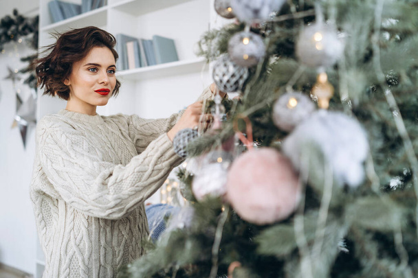 Christmas, New Year. Pretty and happy brunette in warm sweater decorating christmas tree at home - Foto, imagen