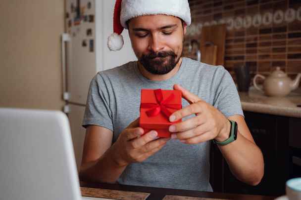 クリスマスの時間に自宅のソファに座って赤いカップを持つヒップスター男性。仮想クリスマスハウスパーティー。自宅からのビデオ会議の呼び出しを満たすオンラインチーム。サンタの帽子をかぶった男 - 写真・画像