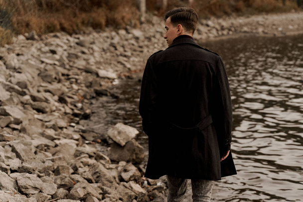 Уверенная мужская модель в черном модном пальто. Красавчик гуляет возле озера - Фото, изображение