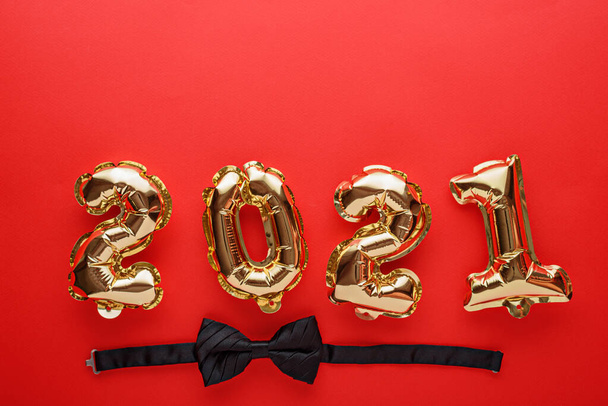 Goldfolie Luftballons Zahlen 2021 mit schwarzer Fliege auf rotem Hintergrund, Neujahrskomposition, festliches Dinner-Konzept, Weihnachtsfeier, Draufsicht, Kopierraum - Foto, Bild