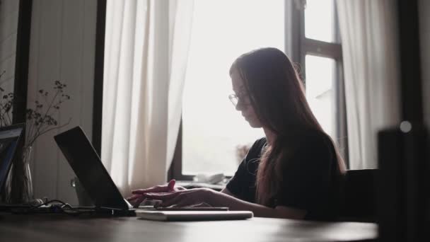 Egy fiatal férfi és egy fiatal lány laptopok előtt dolgoznak a fényes lakásukban. Dolgozz otthonról a karantén alatt. Szabadúszók távoli munkája. Gyönyörű fiatal gondolkodó lány szemüvegben egy számítógép monitor előtt. - Felvétel, videó