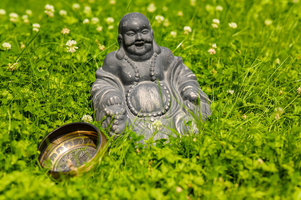 Φιγούρα του Βούδα δίπλα σε ένα μπολ τραγούδι στέκεται στον κήπο στη μέση της άνθισης τριφύλλι. Η εστίαση είναι στο σχήμα. - Φωτογραφία, εικόνα