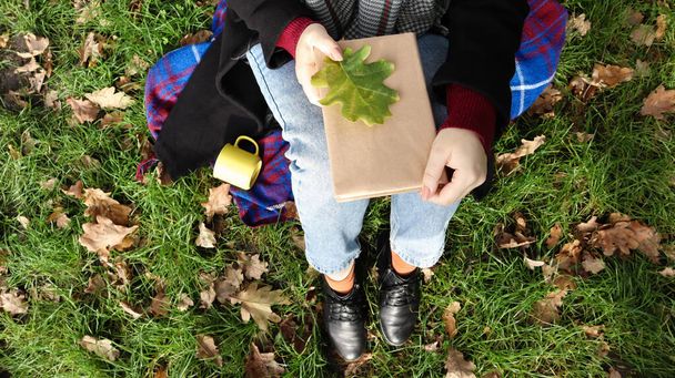 A woman holds a closed book lying on her lap with a fallen oak leaf in a park on a sunny warm autumn day on a green meadow. The concept of relaxation, reading and relaxation alone. Top view, flat lay - Fotó, kép