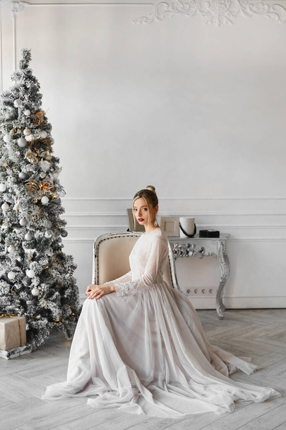 A gorgeous model girl in a fashionable dress sits near the Christmas tree in the vintage interior - Photo, image