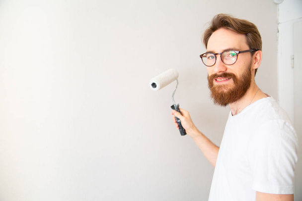 Young man painting wall in white with a paint roller in a bright room. Self apartment renovation, house repair works, walls painting - a house painter at work, looking at camera, with a copy-space. - Photo, Image