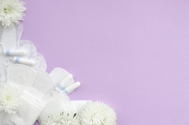 Menstruációs párnák és tamponok lágy fehér virágokkal pasztell lila háttérrel. Női kritikus napok, nőgyógyászati menstruációs ciklus. Egészségügyi női higiénia. Lapos fekvésű fénymásoló - Fotó, kép