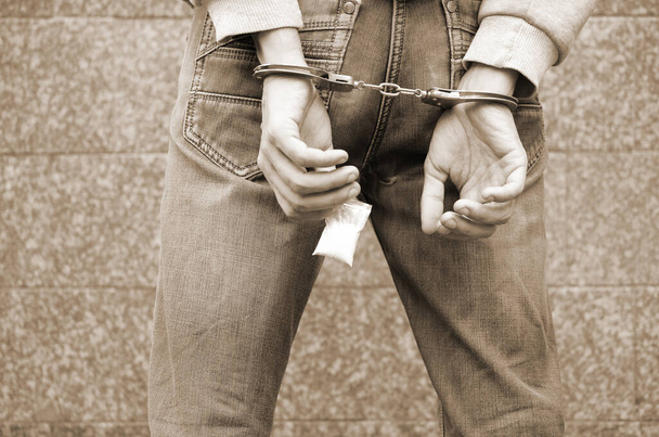 Арестованный наркоторговец в наручниках с маленьким пакетом героина на фоне темной стены. Концепция незаконного оборота наркотиков - Фото, изображение