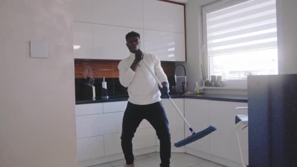Happy black man having fun dancing and singing while cleaning floor in the kitchen. domestic life concept - Footage, Video
