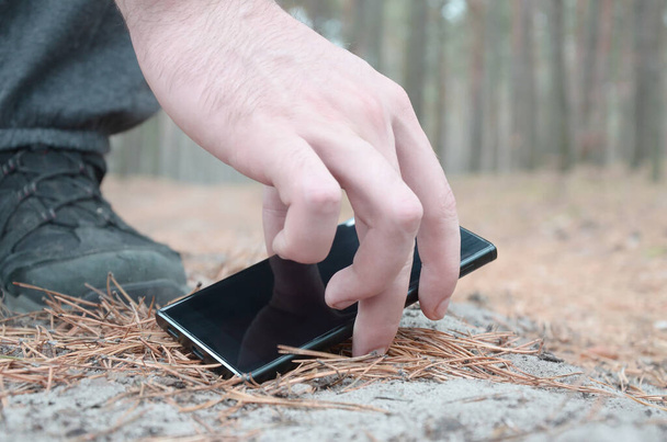 Mano masculina recogiendo un teléfono móvil perdido de un suelo en el camino de madera de abeto de otoño. El concepto de encontrar una cosa valiosa y buena suerte - Foto, imagen