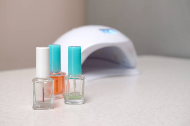 Lámpara de diodo UV para uñas y juego de esmalte de uñas cosmético para manicura y pedicura sobre fondo de mesa blanco. Concepto de procedimientos de belleza para mujeres. Frascos transparentes - Foto, Imagen