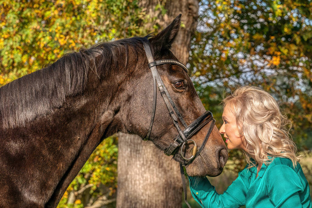 Fiatal szőke nő zöld ruhában, egy barna lóval csókolózik. Az erdőben ősszel arany színben.. - Fotó, kép