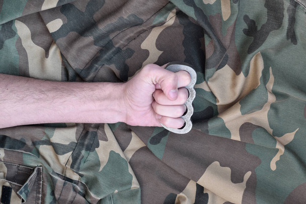 Мужской кулак с медными костяшками на фоне камуфляжной куртки. Концепция скинхедов, домашнее оружие ближнего боя - Фото, изображение