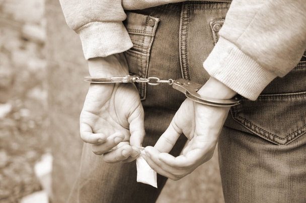 Gearresteerde drugsdealer in handboeien met kleine heroïne drugspakket op donkere muur achtergrond. Illegale drugshandel - Foto, afbeelding