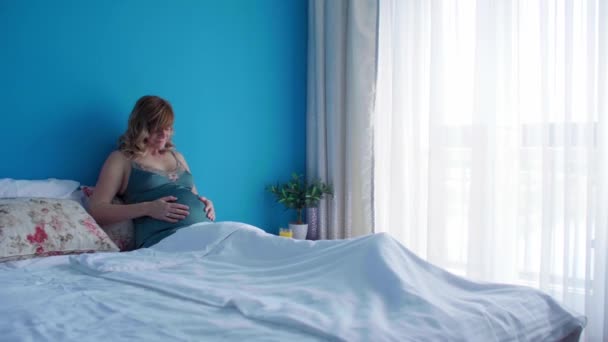 Una mujer embarazada sonriente en una posición semi-sentada en la cama con las manos alrededor del estómago. Está mirando hacia su estómago.. - Imágenes, Vídeo