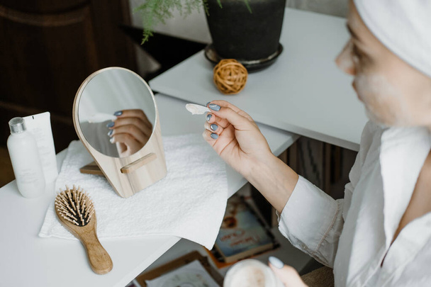 Jonge vrouw in witte handdoek chillen in de slaapkamer en het maken van klei gezichtsmasker in de buurt van spiegel. Meisje doet schoonheidsbehandelingen en ontspant thuis. Ochtend huidverzorging schoonheid routine, zelfverzorging - Foto, afbeelding