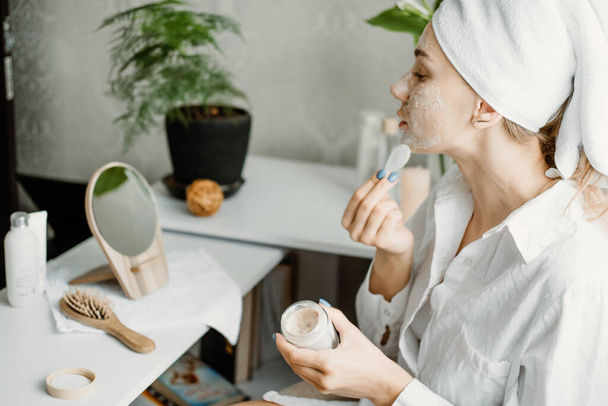 Young woman in white towel chilling in bedroom and making clay facial mask near mirror. Girl doing beauty treatment and relaxing at home. Morning skin care beauty routine, self care - Photo, Image