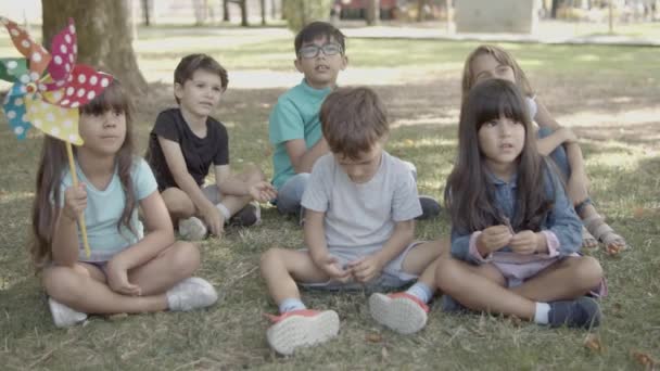 Niños multiétnicos sentados en el césped en el parque de la ciudad juntos - Imágenes, Vídeo