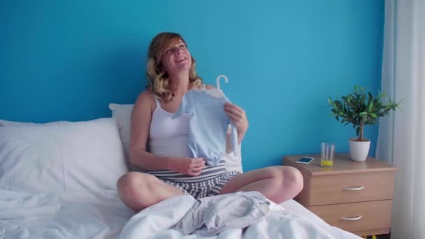 Uma mulher branca grávida sorridente e feliz em uma posição sentada em sua cama enquanto olha para roupas de bebê minúsculas. - Filmagem, Vídeo