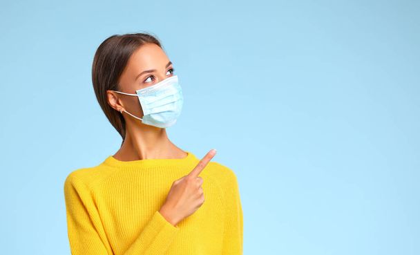 Orvosi maszkos nő, aki a válla fölött lévő üres helyre mutat a koronavírus-járvány idején a kék háttér ellen. - Fotó, kép