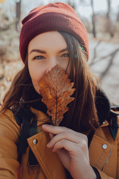 young brunette woman in maroon knitted hat, yellow leather jacket and black hoodie holds yellow dry oak leaf to her face and smiles, vertical unfiltered aesthetic atypical portrait - Photo, Image