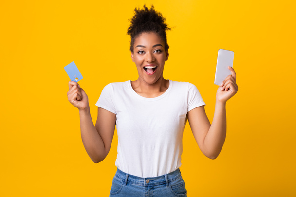 Excited black lady holding credit card and cellphone at studio - Фото, изображение