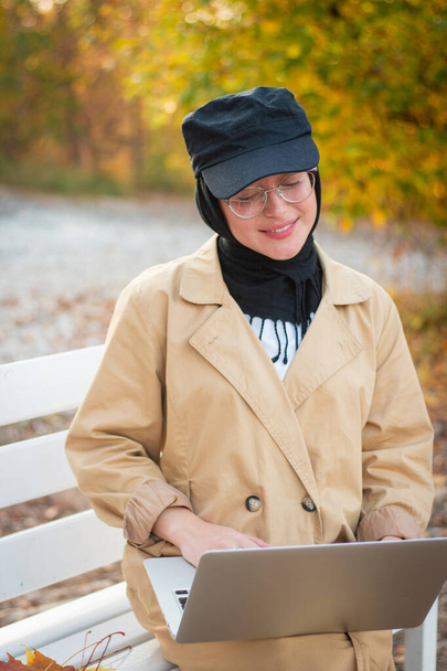 A Muslim woman in a hijab in a beige trench coat sits on a bench with a cup of coffee and a laptop in the park. Muslim business woman. Student. Glasses for sight. - Фото, изображение