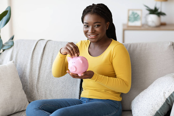 幸せなアフリカ系アメリカ人女性が貯金箱を持ち、自宅でソファに座って - 写真・画像
