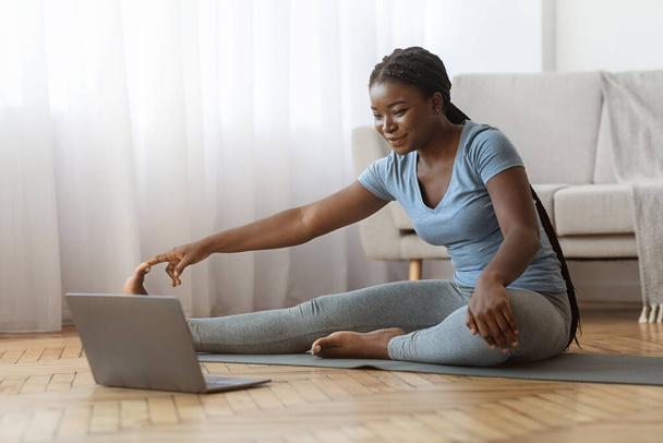 Entrenamiento en casa. Deportiva mujer africana estirando los músculos de las piernas delante de la computadora portátil - Foto, Imagen