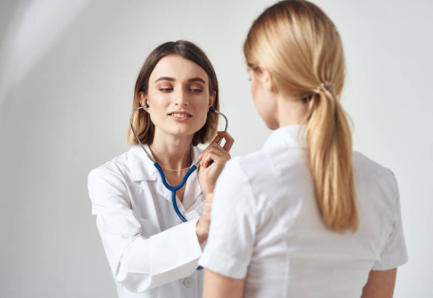 médecin avec un stéthoscope examine une femme dans un t-shirt blanc sur un fond clair - Photo, image