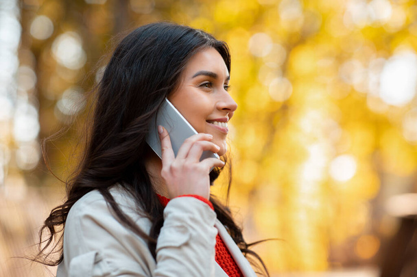 Jovencita positiva hablando por celular en el parque de otoño en un día soleado brillante, espacio para copiar - Foto, imagen