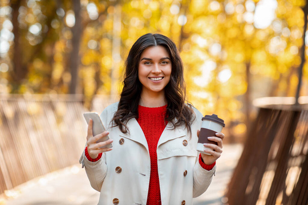 Portret van een mooie dame met koffie te gaan en smartphone glimlachen en kijken naar de camera buiten op koele herfstdag - Foto, afbeelding