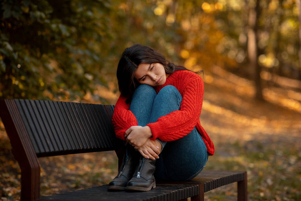 Depresyonda olan genç bir kadın, sarı sonbahar parkında bankta otururken dizlerine sarılıyor. - Fotoğraf, Görsel