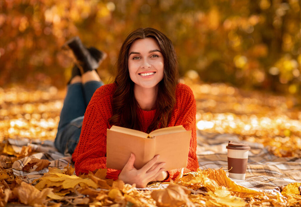 Jovem sorridente com livro e café para ir deitado em cobertor entre folhas de outono caídas e desfrutando de dia quente de outono - Foto, Imagem