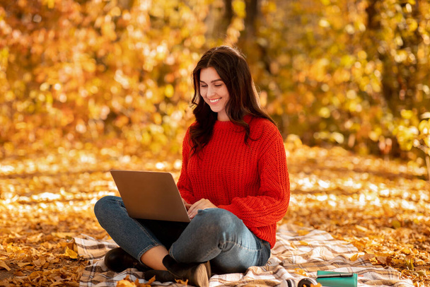 Милая белая женщина использует ноутбук для онлайн-работы, просматривая веб-сайты в городском парке осенью - Фото, изображение