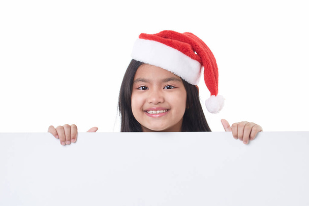 Portret szczęśliwej dziewczynki w kapeluszu Mikołaja pozowanie za białym panelem izolowane na białym tle trzymając pusty plansza Boże Narodzenie billboard - Zdjęcie, obraz