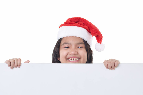 Muotokuva onnellinen pieni tyttö yllään Santa hattu poseeraa takana valkoinen paneeli eristetty valkoisella pohjalla tilalla tyhjä aluksella joulun mainostaulu - Valokuva, kuva