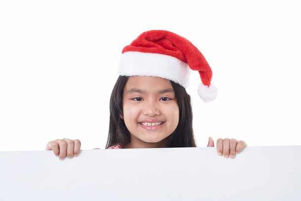 Porträt eines glücklichen kleinen Mädchens mit Weihnachtsmütze posiert hinter einem weißen Panel isoliert auf weißem Hintergrund hält eine weiße Tafel Weihnachten Plakatwand - Foto, Bild