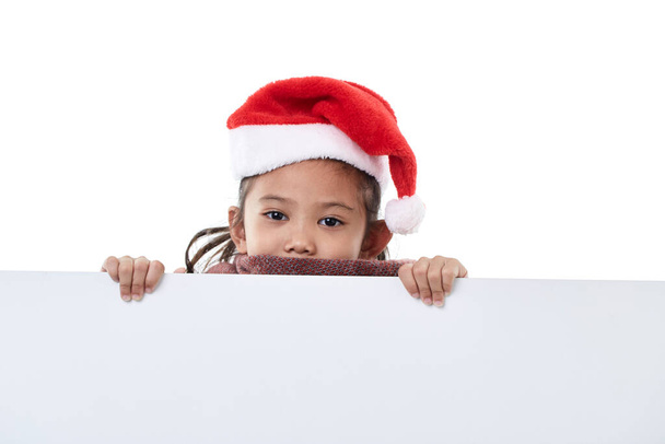 Noel Baba şapkası takan mutlu küçük bir kızın portresi beyaz arka planda izole edilmiş beyaz bir panonun arkasında boş bir Noel panosu tutuyor. - Fotoğraf, Görsel
