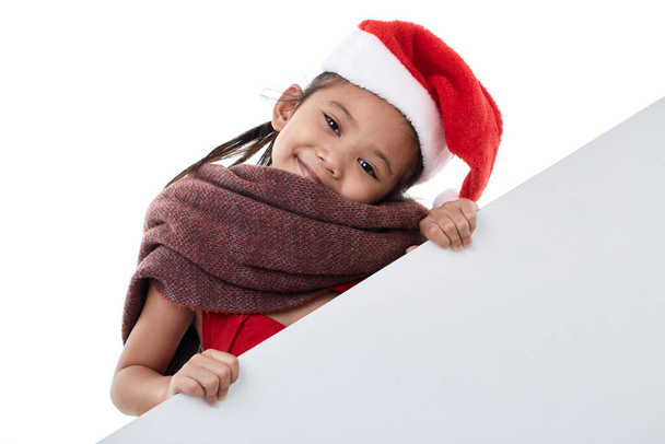 Ritratto di una bambina felice che indossa il cappello di Babbo Natale posando dietro un pannello bianco isolato su sfondo bianco con un cartellone bianco - Foto, immagini