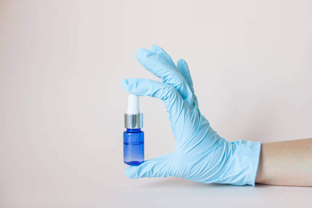 Bottles with serum for the face in his hands in protective medical rubber gloves on a light background. Beauty industry. - Фото, изображение