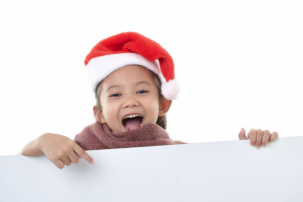 幸せな女の子の肖像画を身に着けているサンタの帽子白い背景に隔離された白いパネルを指す空白の板クリスマスの看板 - 写真・画像