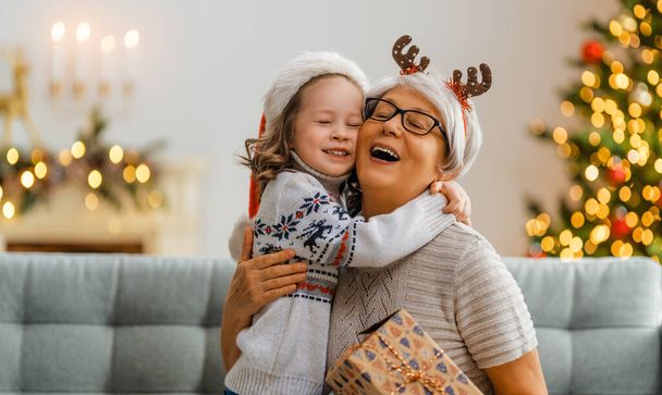 Merry Christmas and Happy Holidays! Cheerful grandma and her cute grand daughter girl exchanging gifts. Granny and little child having fun near tree indoors. Loving family with presents in room. - Photo, Image