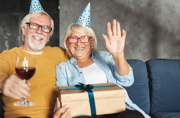 Happy senior couple sitting at home on the sofa with gift have video call look at camera - Foto, Imagem