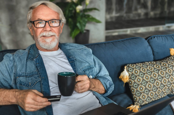 Portrait of an elderly man with a credit card and a cup of coffee sitting on the couch at home. Shopping online with credit card concept - Photo, Image