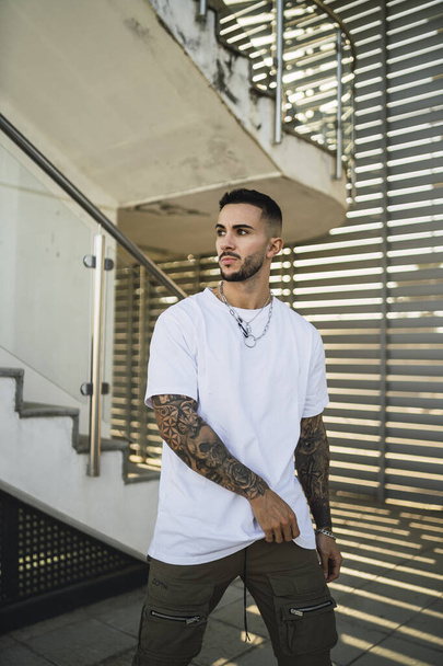 A sexy tattooed man wearing a white shirt and joggers standing on the background of a staircase - Foto, immagini